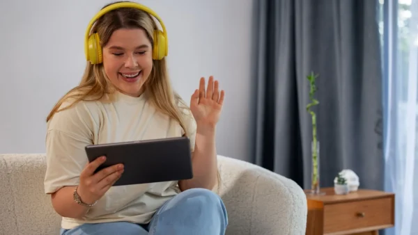 Mulher jovem sorrindo no sofá, usando fones de ouvido over-ear amarelos e acenando em uma videochamada no tablet